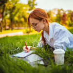 female-student-prepares-exams-using-laptop-makes-notes-lying-grass-education-concept